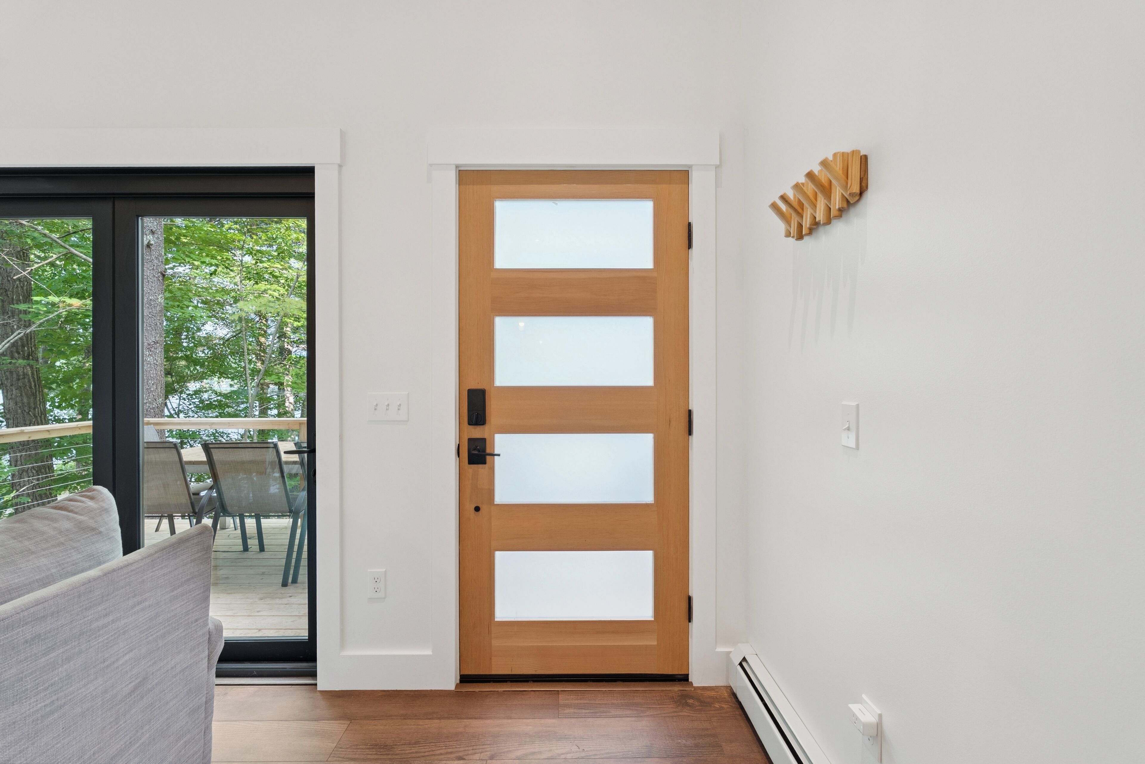 A modern interior with a wooden door, a glass door leading to an outdoor area, and a white wall with a wooden art piece.