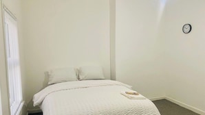 Ein Schlafzimmer mit einem Einzelbett, weißer Bettwäsche, einer Wanduhr und einem Fenster mit Jalousien.