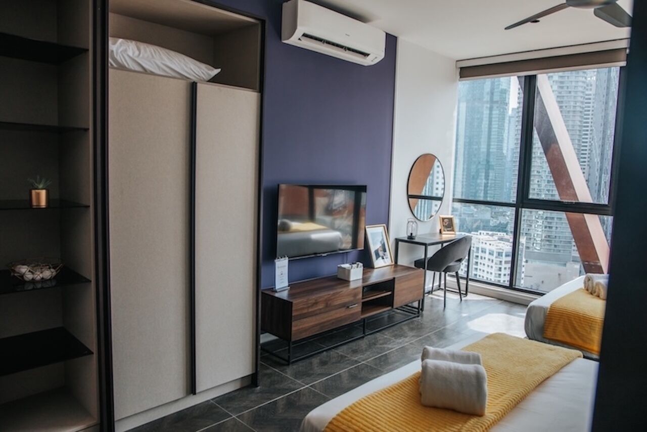 A modern hotel room with a bed, a desk, a TV, and a large window offering a city view.