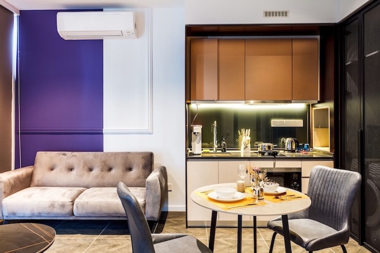 A modern living room with a sofa and a dining table set for two, a kitchen with wooden cabinets, and a large window.