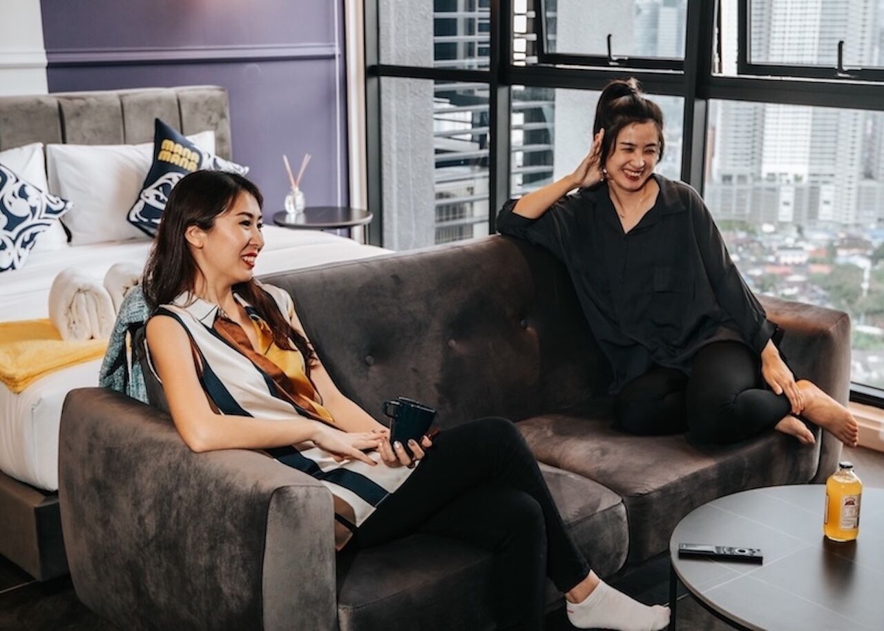 Two people sitting on a grey sofa in a modern room with a city view through large windows.