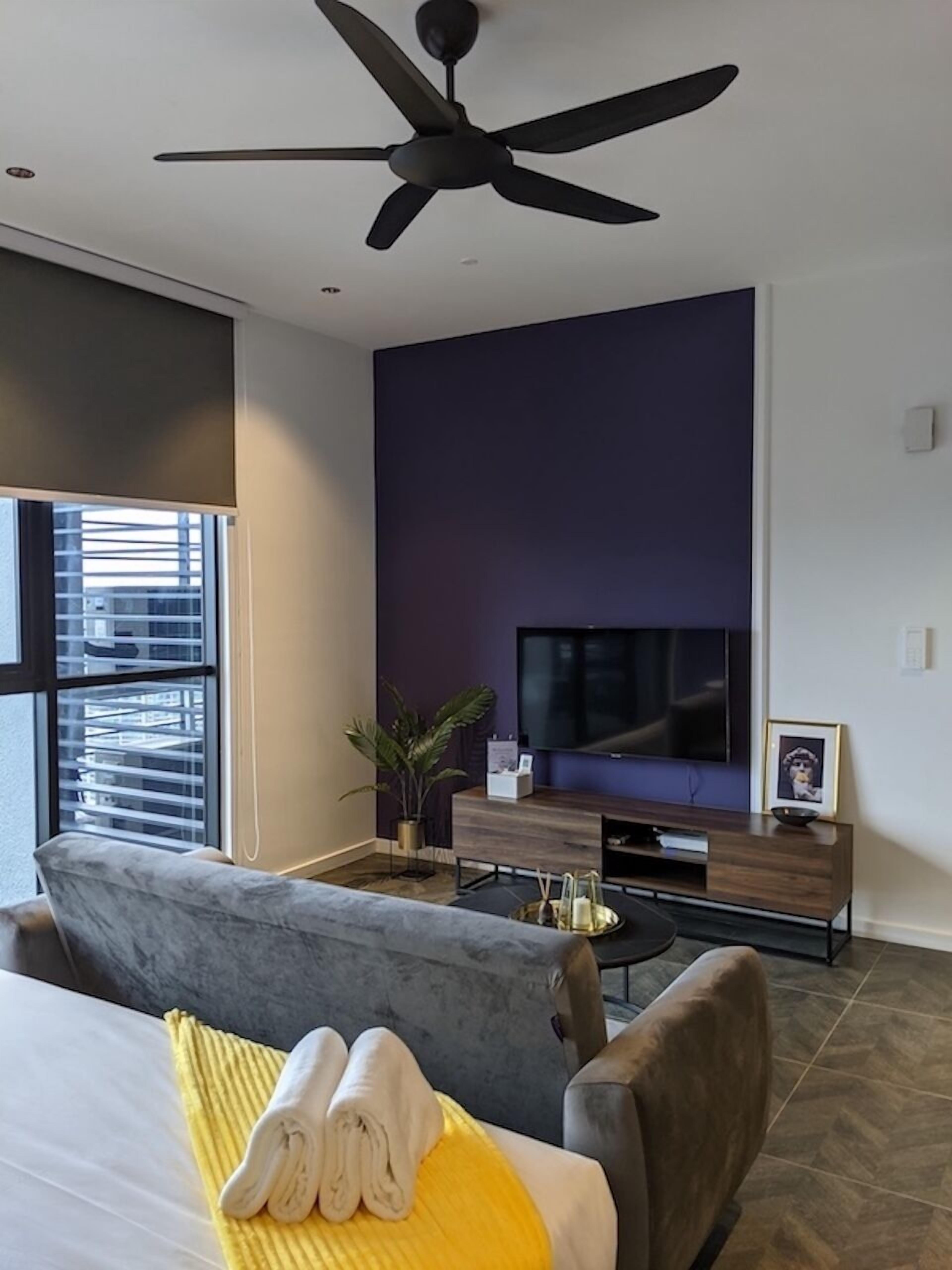 Ein modernes Hotelzimmer mit einem großen Bett, einem grauen Sofa, einem Deckenventilator, einem Fernseher auf einem Holzboden und einem Fenster mit Jalousien.