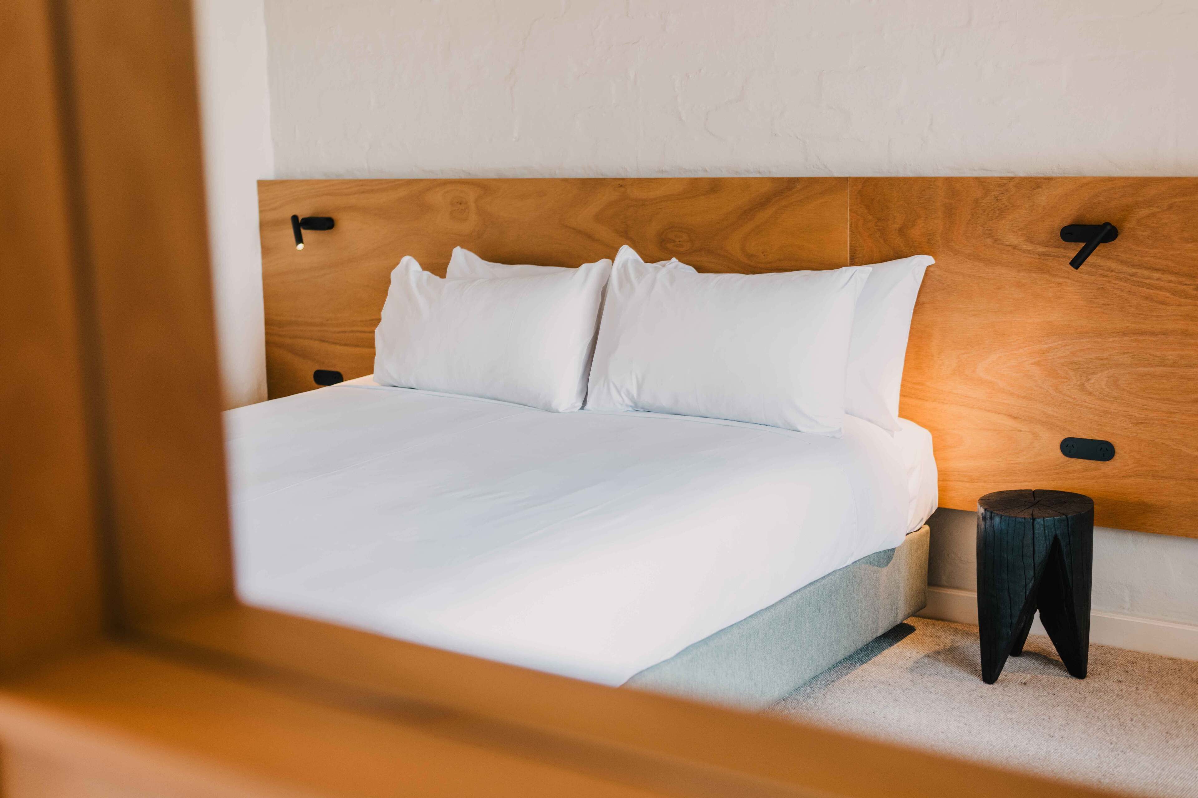 Een netjes opgemaakt bed met witte lakens, een houten hoofdbord en een klein nachtkastje ernaast.