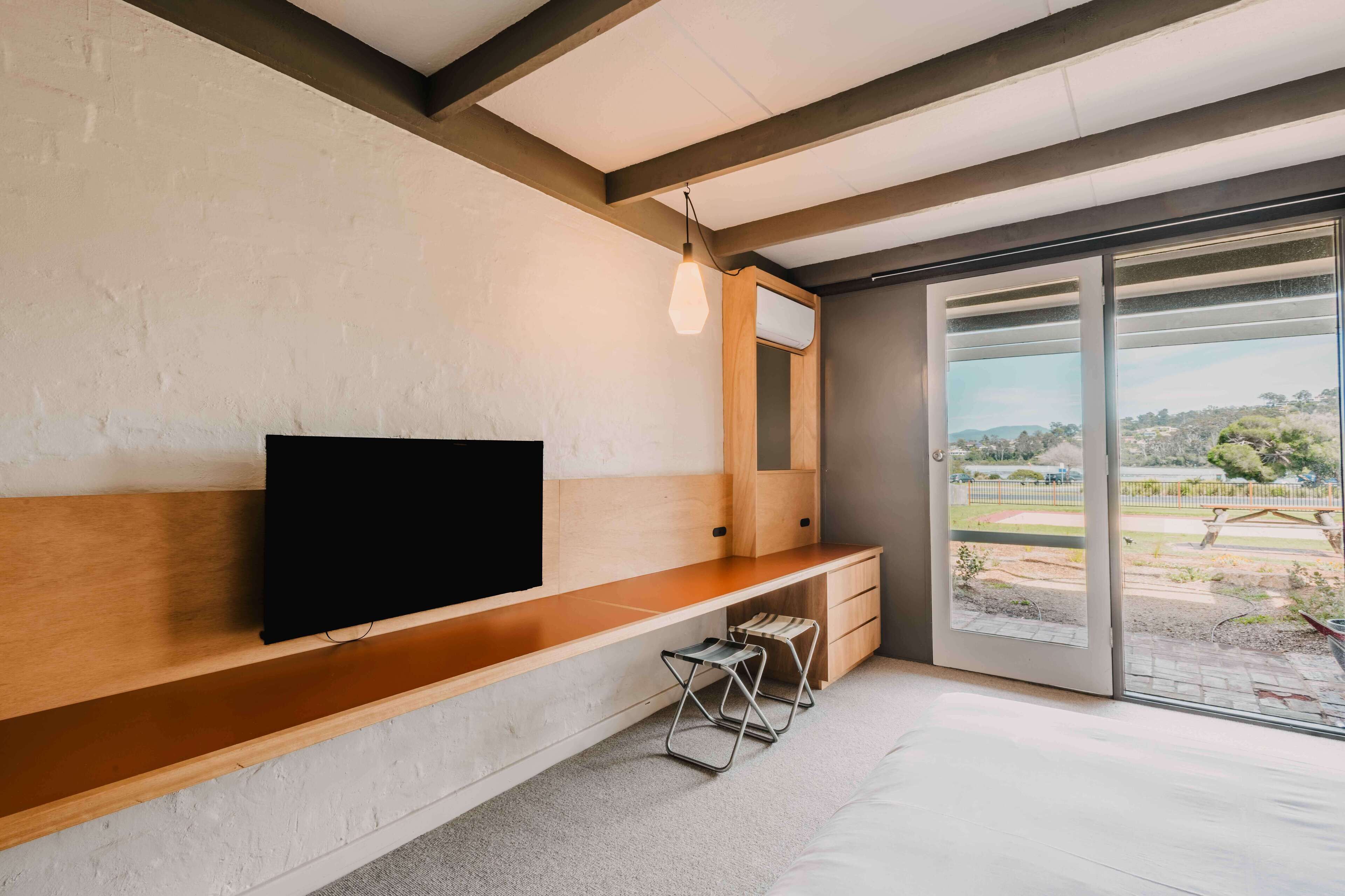 Una habitación moderna con un escritorio de madera, un televisor de pantalla plana, una lámpara colgante y un ventanal corredero que da a un espacio exterior.