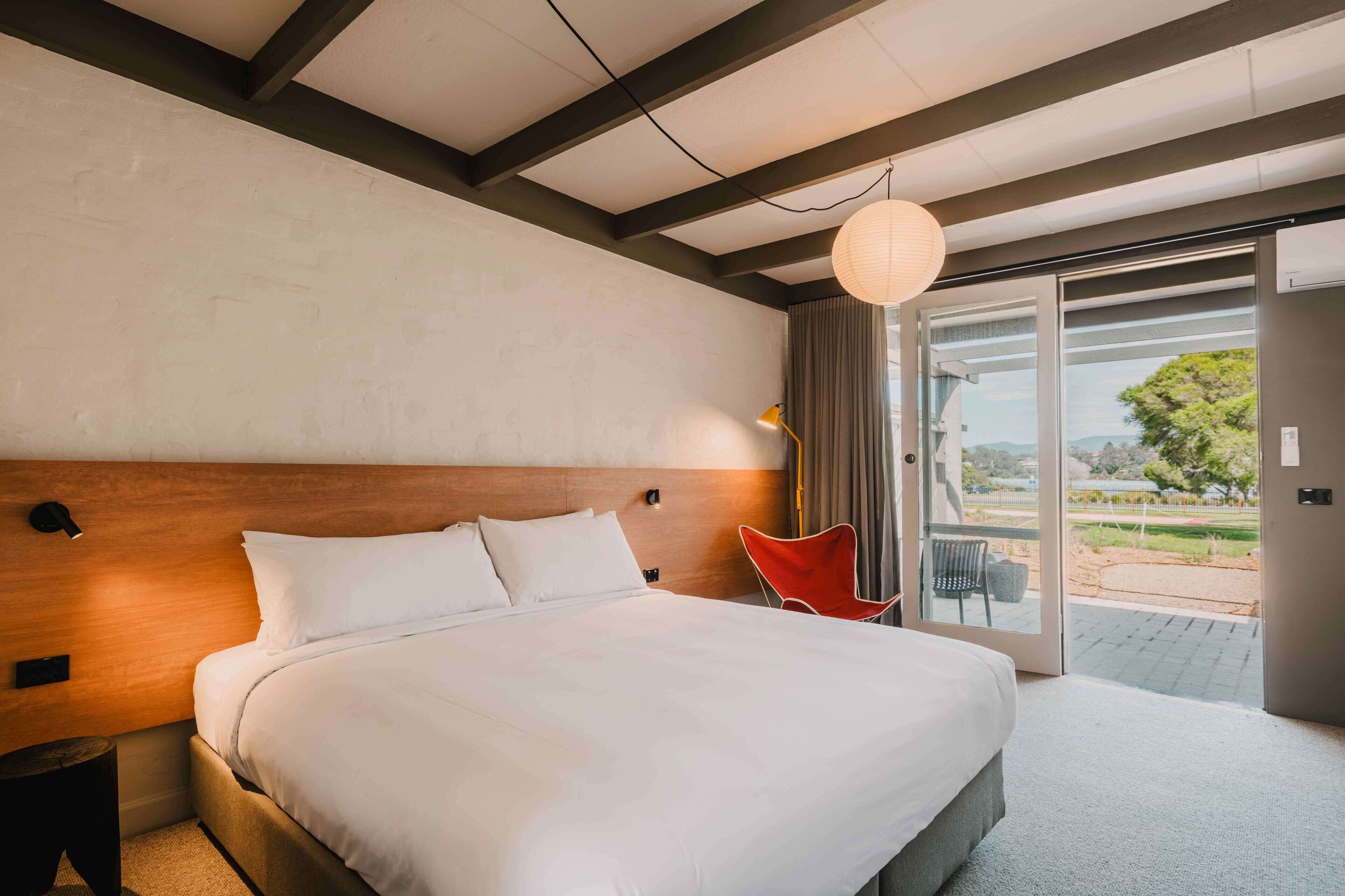 Ein Hotelzimmer mit einem großen Bett, einem Holzkopfbrett, einem Nachttisch, einem roten Sessel und Blick ins Freie durch eine Glastür.