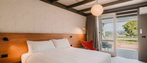 A hotel room with a large bed, a wooden headboard, a nightstand, a red chair, and a view of the outdoors through a glass door.
