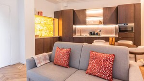 Een moderne woonkamer met een grijze bank en rode kussens met een patroon, een keuken met houten kastjes en roestvrijstalen apparatuur, en een eethoek met een ronde tafel en stoelen.