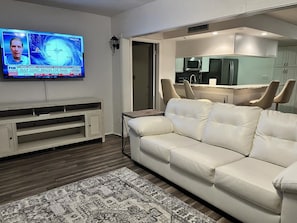 A living room with a flat-screen TV mounted on the wall displaying a weather forecast, a white leather sofa, a wooden entertainment unit, and a kitchen area with modern appliances. - Peaceful Condo -Golf Course & Sunset Views -Min stay required (Fort myers)