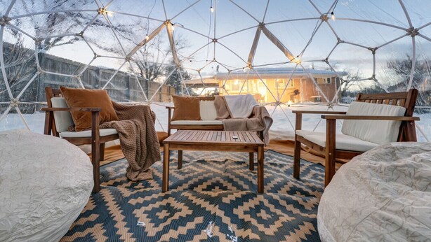 Interieur van een geodetische koepel met houten meubels, een vloerkleed met patroon en lichtslingers.