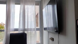 Et værelse med stort vindue, lette gardiner, et fladskærms-tv monteret på væggen, en mørk lænestol og et lille bord med billedramme og en dekorativ genstand.