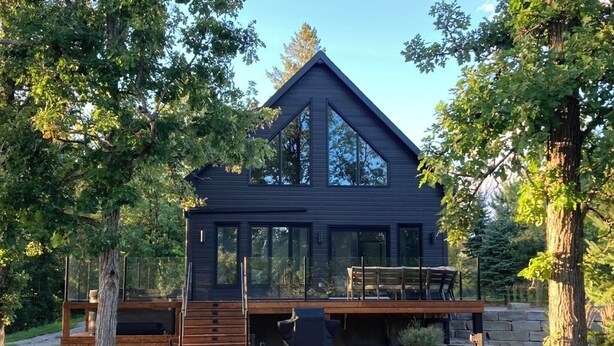 A modern wooden cabin with a large glass window, a deck, and a stone wall.