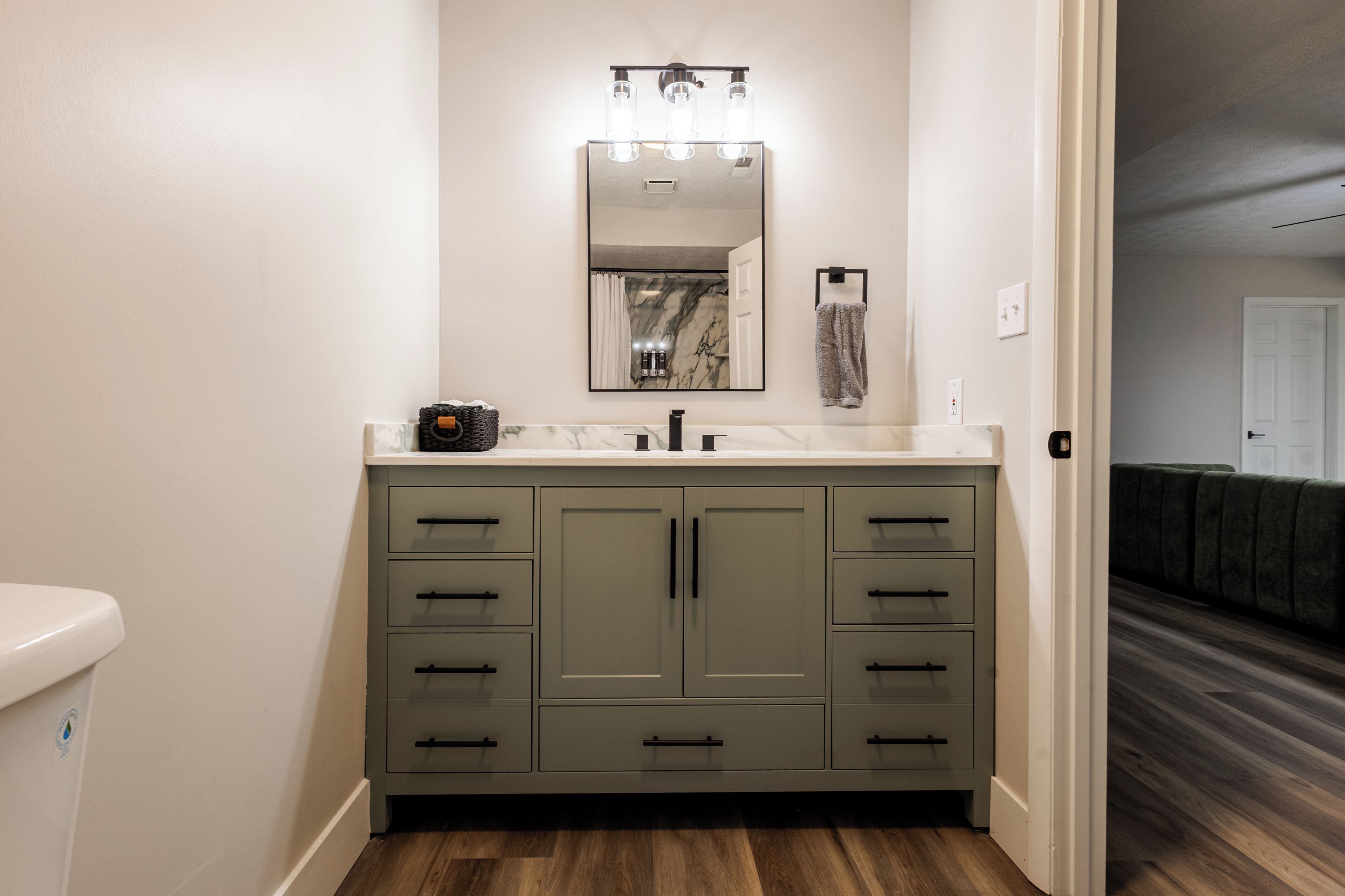 A bathroom with a marble countertop, a sink, a mirror with lights, and a green cabinet.