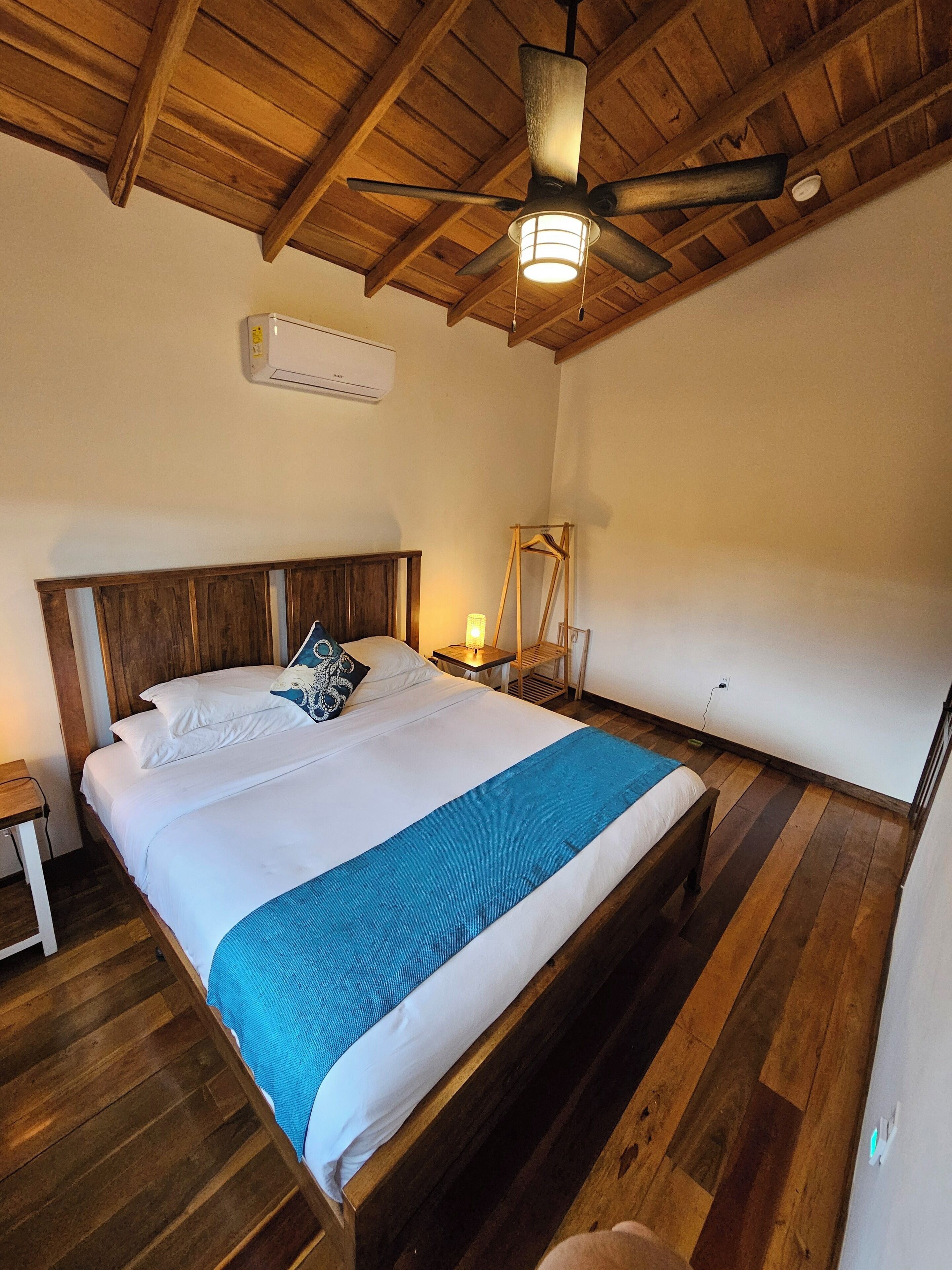 Un dormitorio con techo de madera, cama con ropa de cama blanca y una manta azul, mesita de noche con lámpara, y lámpara colgante.