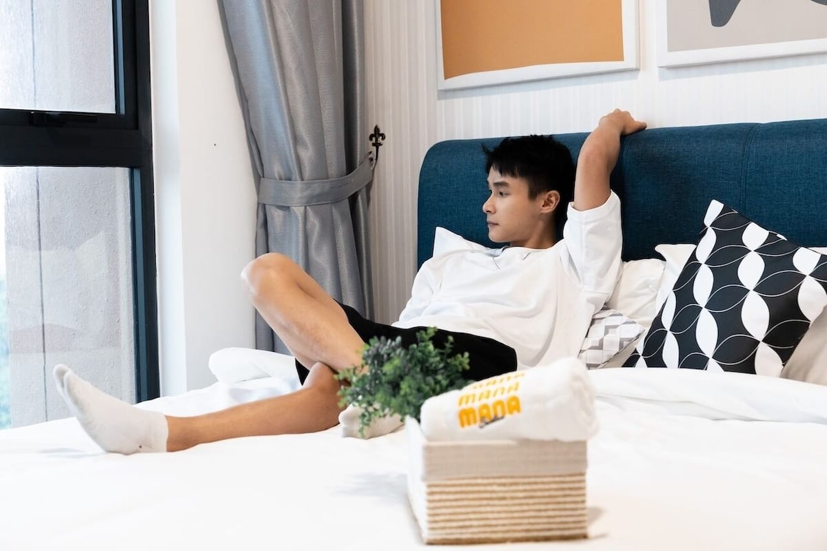 A person lying on a bed with a blue headboard, white bedding, and a black and white patterned pillow.