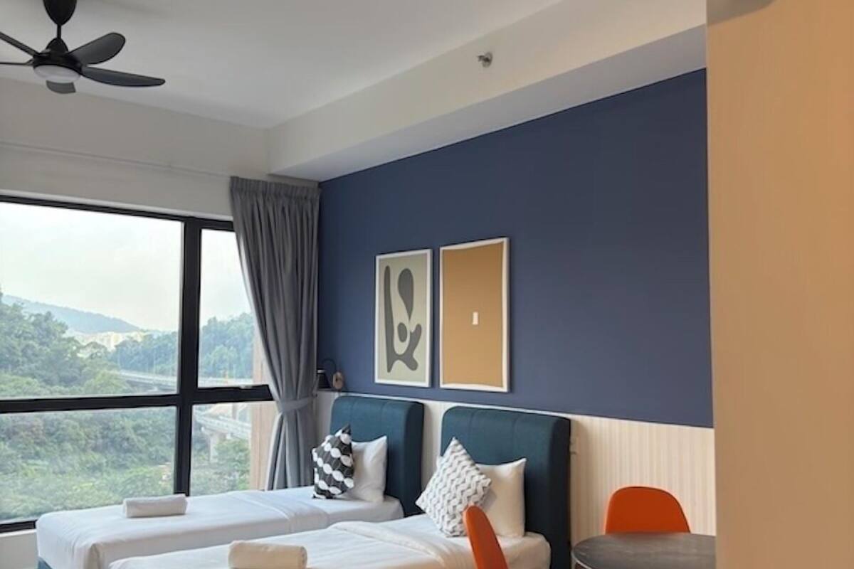 A hotel room with two beds, a dining table, and chairs. There is a ceiling fan, a window with a view of greenery, and a wall-mounted air purifier.