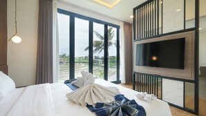 A hotel room with a large bed, a flat-screen TV mounted on the wall, and a view of palm trees and a water body outside. - Bellevue Villa Canggu (Canggu)