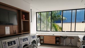 A modern kitchen with built-in appliances, a large window with a view of palm trees, and a grey sofa. - Beautiful studio apartment 400m from Allianz Parque. (São Paulo)