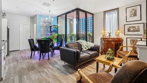 Ein modernes Wohnzimmer mit einer Couch, Sesseln, einem Couchtisch und einem Fernseher. Eine Glaswand trennt den Wohnbereich von einem Balkon mit Pflanzen und Blick auf die Stadt.
