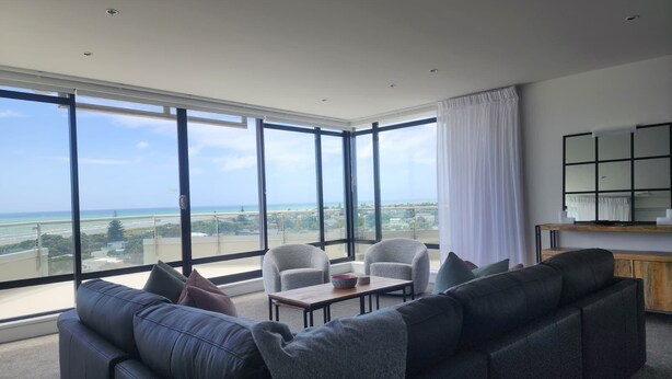 Una sala de estar moderna con un sofá grande, dos sillones, una mesa de centro y vistas al océano y al paisaje urbano a través de ventanales del suelo al techo.