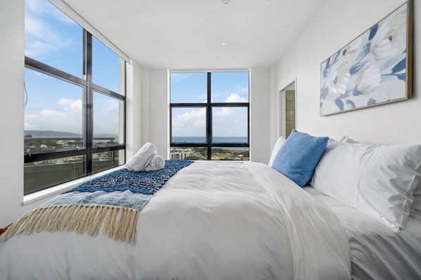A bedroom with a large window, a bed with white and blue bedding, and a blue and beige throw blanket.