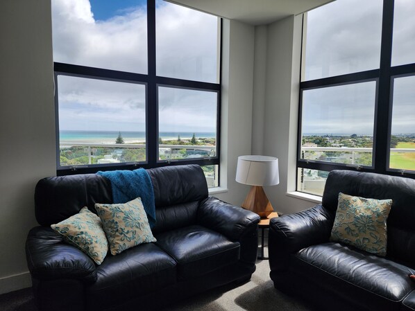 A modern living room with a black leather sofa, two matching armchairs, and a wooden side table with a lamp. Large windows provide a view of a coastal landscape.