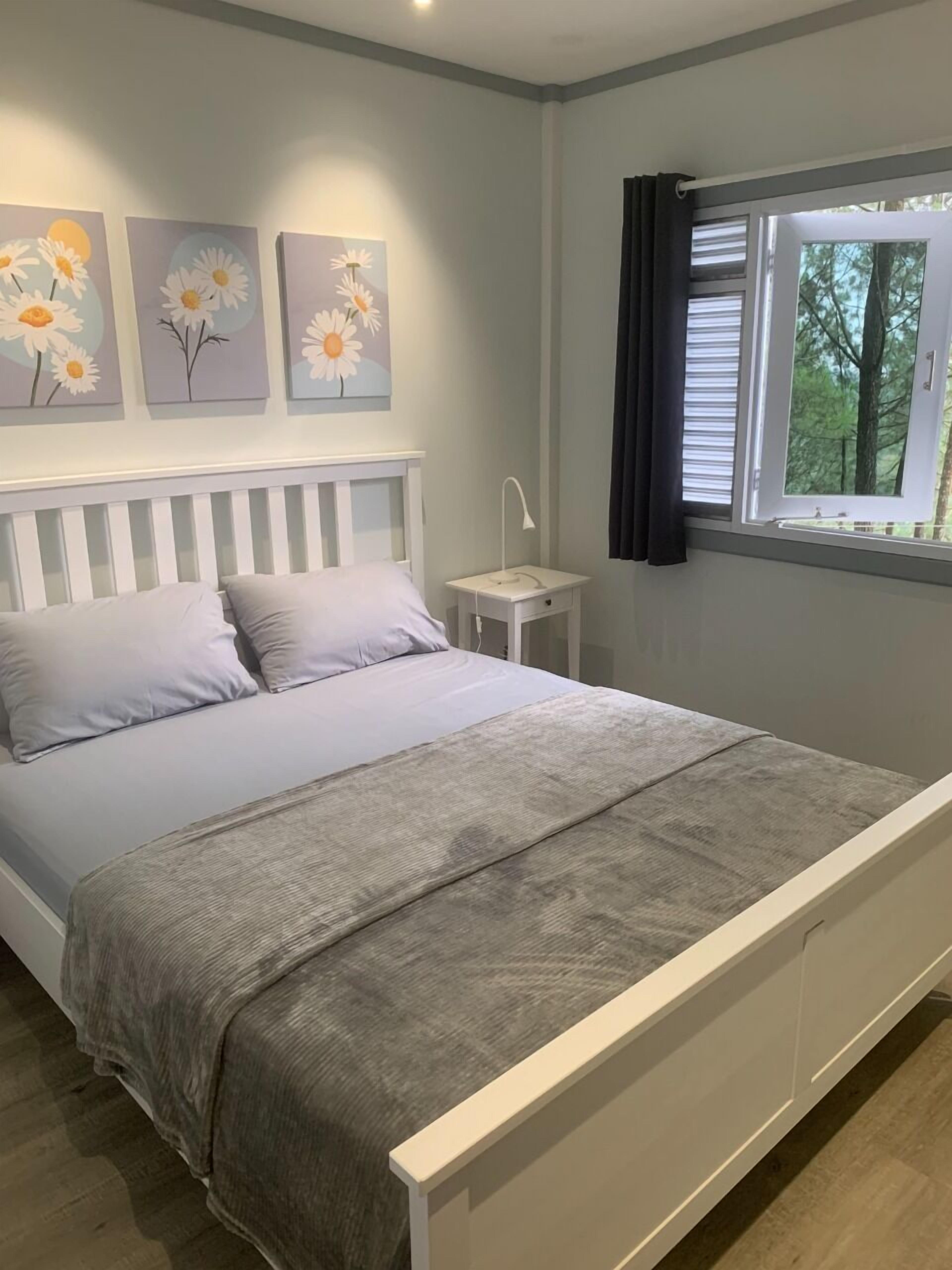 A bedroom with a bed, a nightstand, a window with curtains, and three wall-mounted flower paintings.