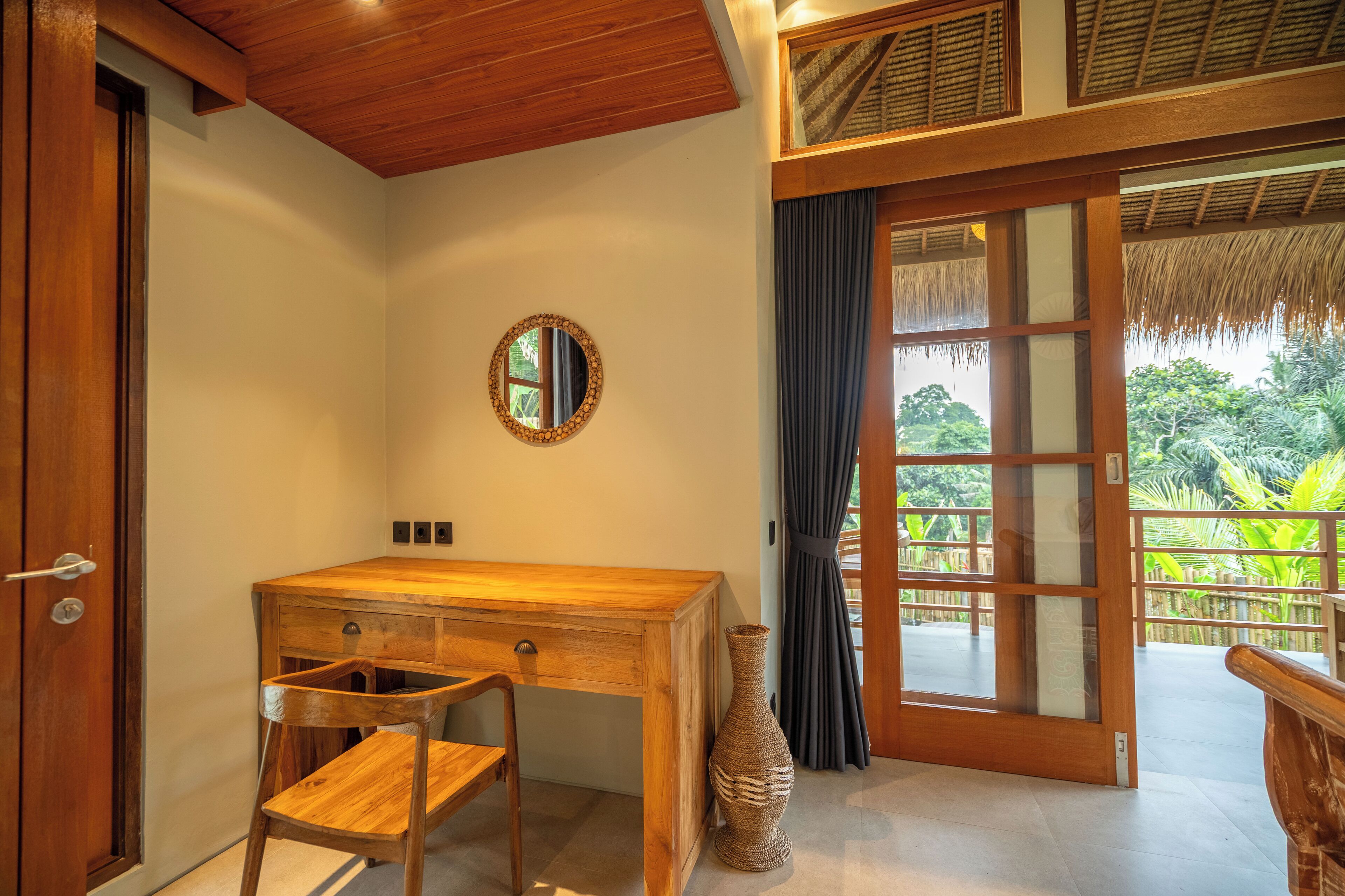 Uma escrivaninha de madeira com uma cadeira, um espelho e um vaso num quarto com uma grande porta de correr de vidro que dá para uma varanda com vista para o verde.