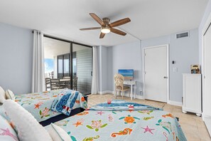 A bedroom with two beds, a ceiling fan, a desk with a chair, a TV, and a sliding glass door leading to a balcony. - Seas The Day- Oceanfront Daytona Beach Getaway – Huge Balcony, Steps to the Sand (Daytona Beach)