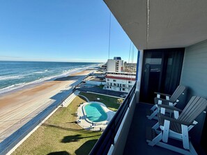 A balcony with two chairs overlooking a beach and a swimming pool. - Seas The Day- Oceanfront Daytona Beach Getaway – Huge Balcony, Steps to the Sand (Daytona Beach)