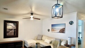 A modern living room with a sofa, a dining table with chairs, a ceiling fan, a decorative hanging light, and a painting on the wall.
