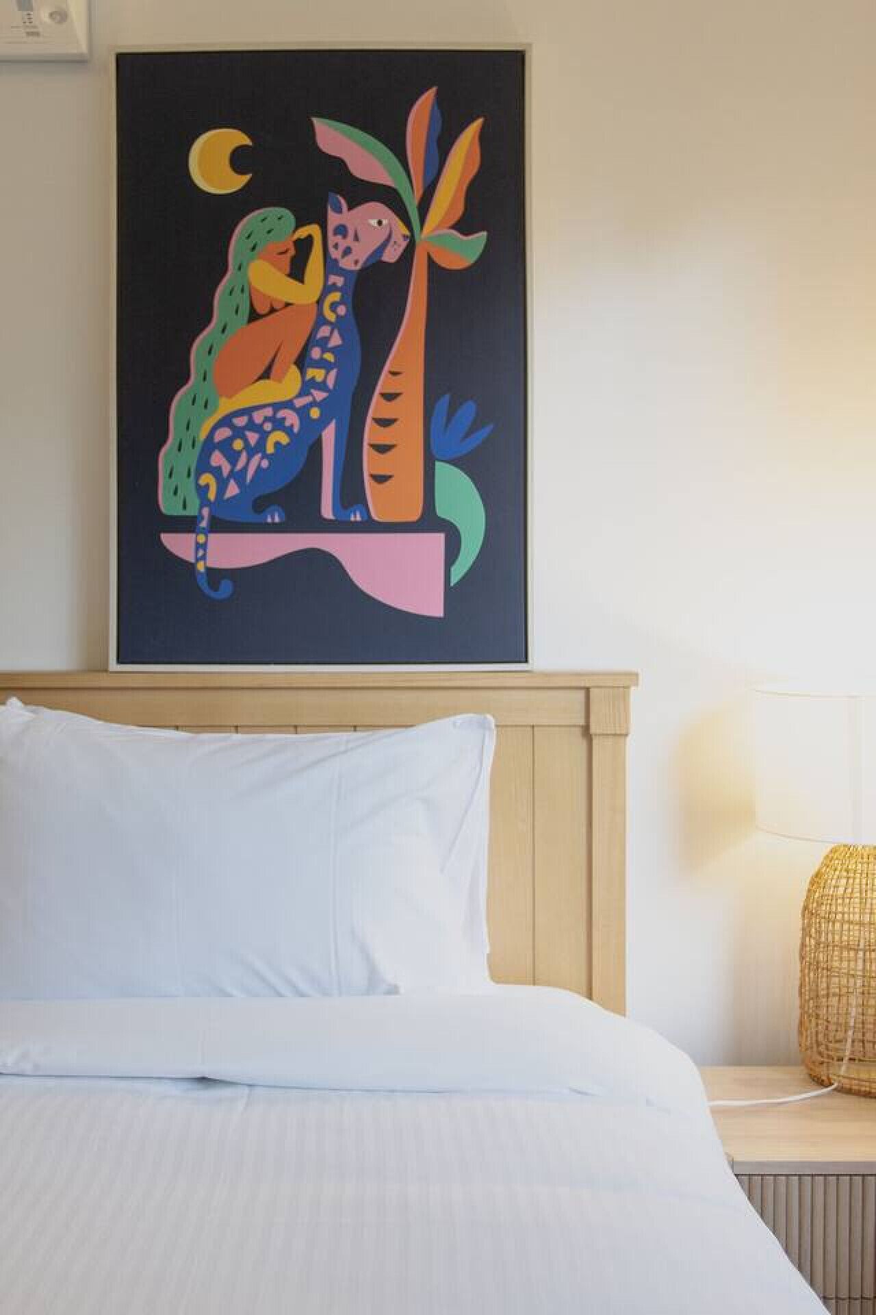 A bed with white linens, a framed abstract painting above it, and a bedside lamp on a wooden nightstand.