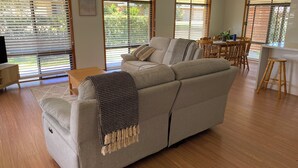 A living room with a sofa, wooden flooring, and large windows with blinds. -  Coastal Palms  Relaxing pet friendly accommodation with pool (Yamba)