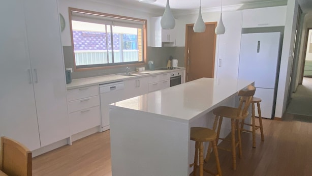 A modern kitchen with a large island, wooden stools, and white cabinetry. -  Coastal Palms  Relaxing pet friendly accommodation with pool (Yamba)