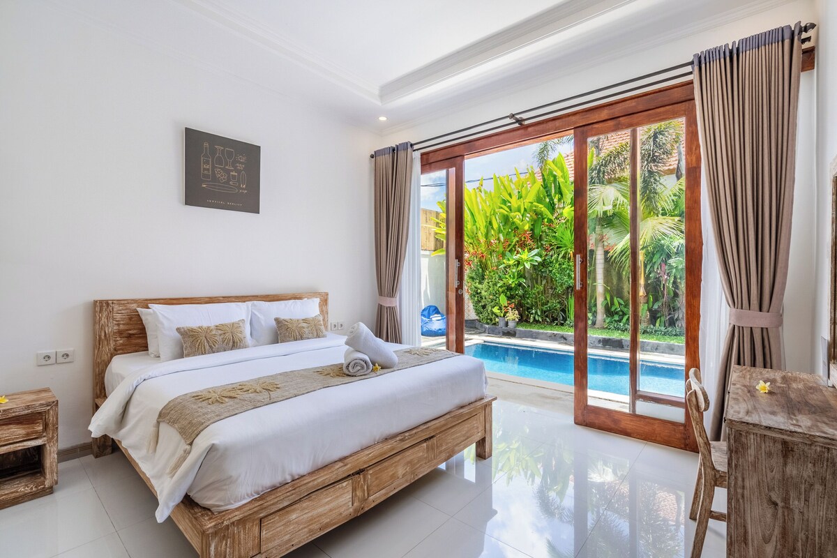 A bedroom with a wooden bed, a view of a swimming pool, and a tropical garden outside.