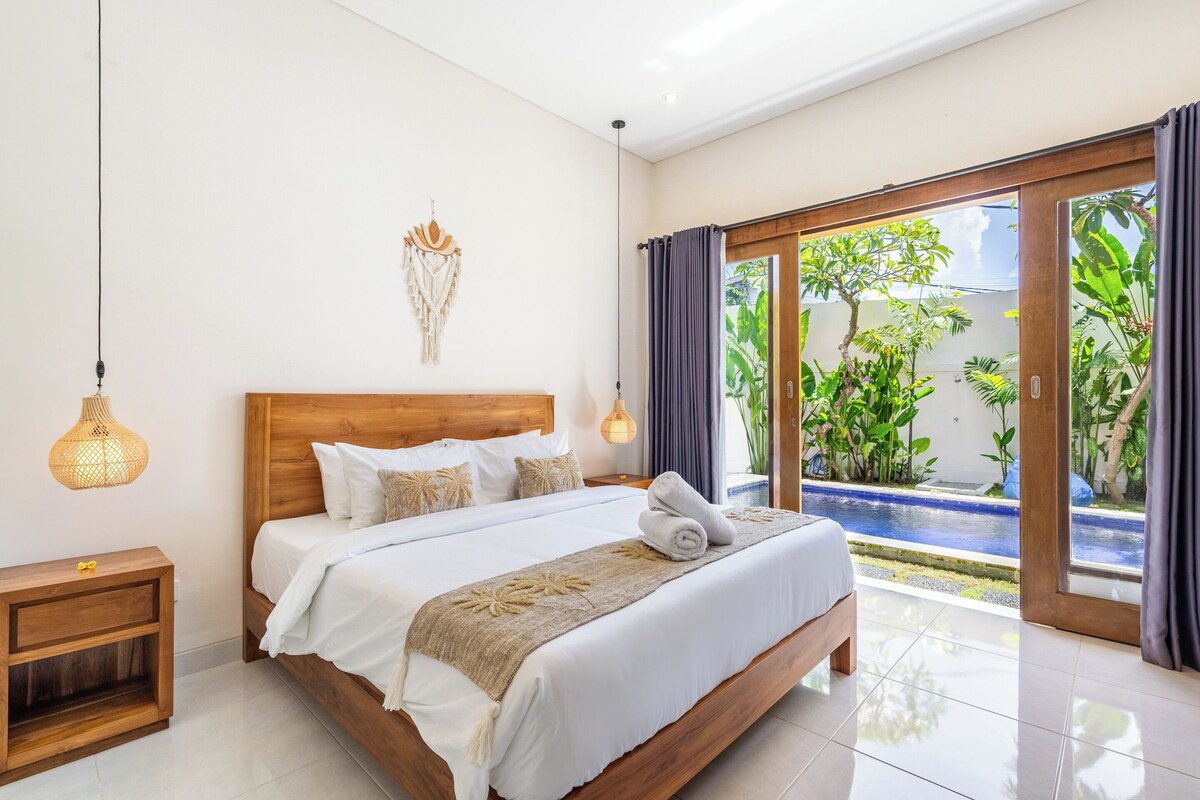 A spacious bedroom with a large bed, a wooden headboard, a bedside table, and a view of a garden through a sliding glass door.
