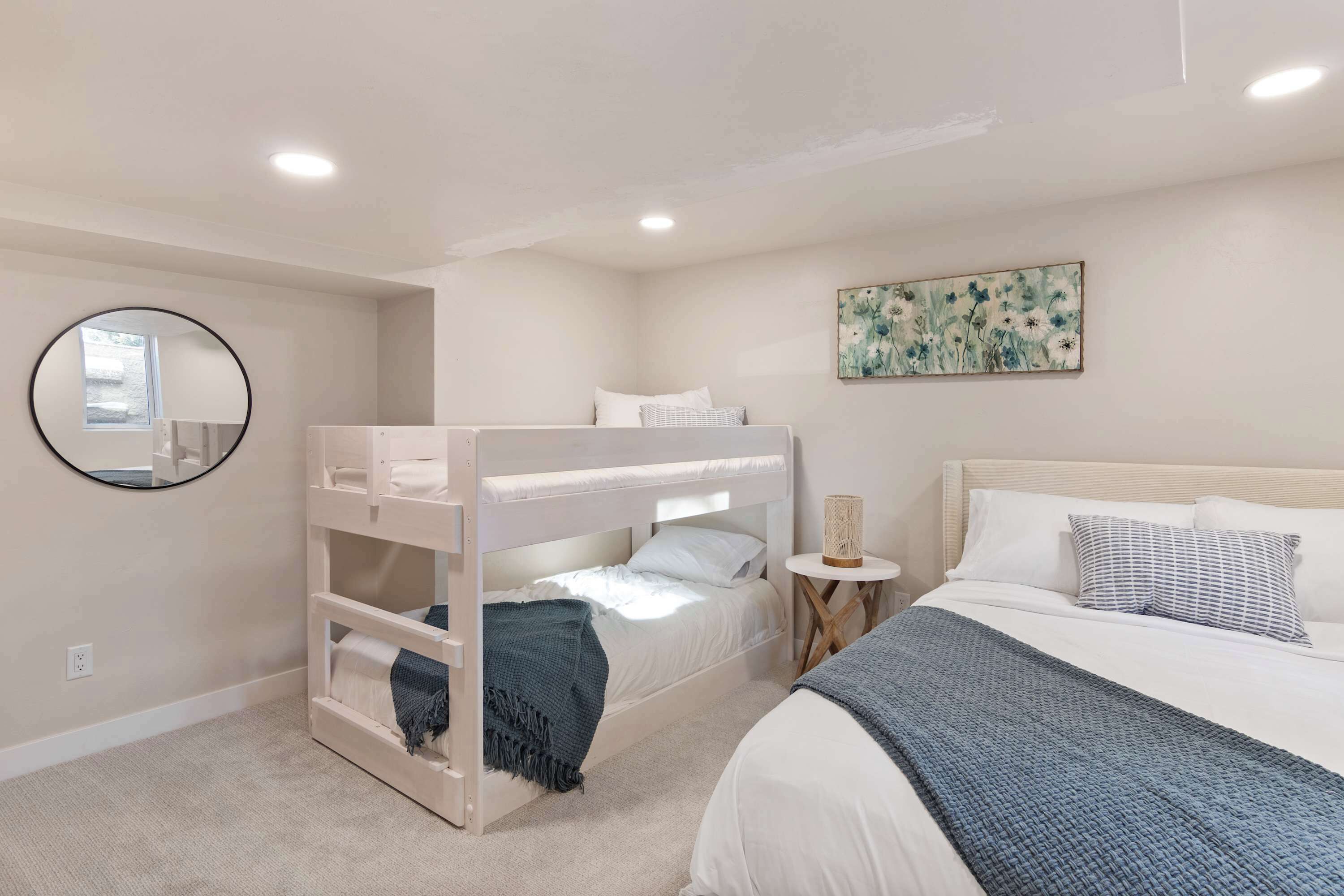 Un dormitorio con literas, un espejo redondo, una mesita de noche con una lámpara y un cuadro con motivos florales en la pared.