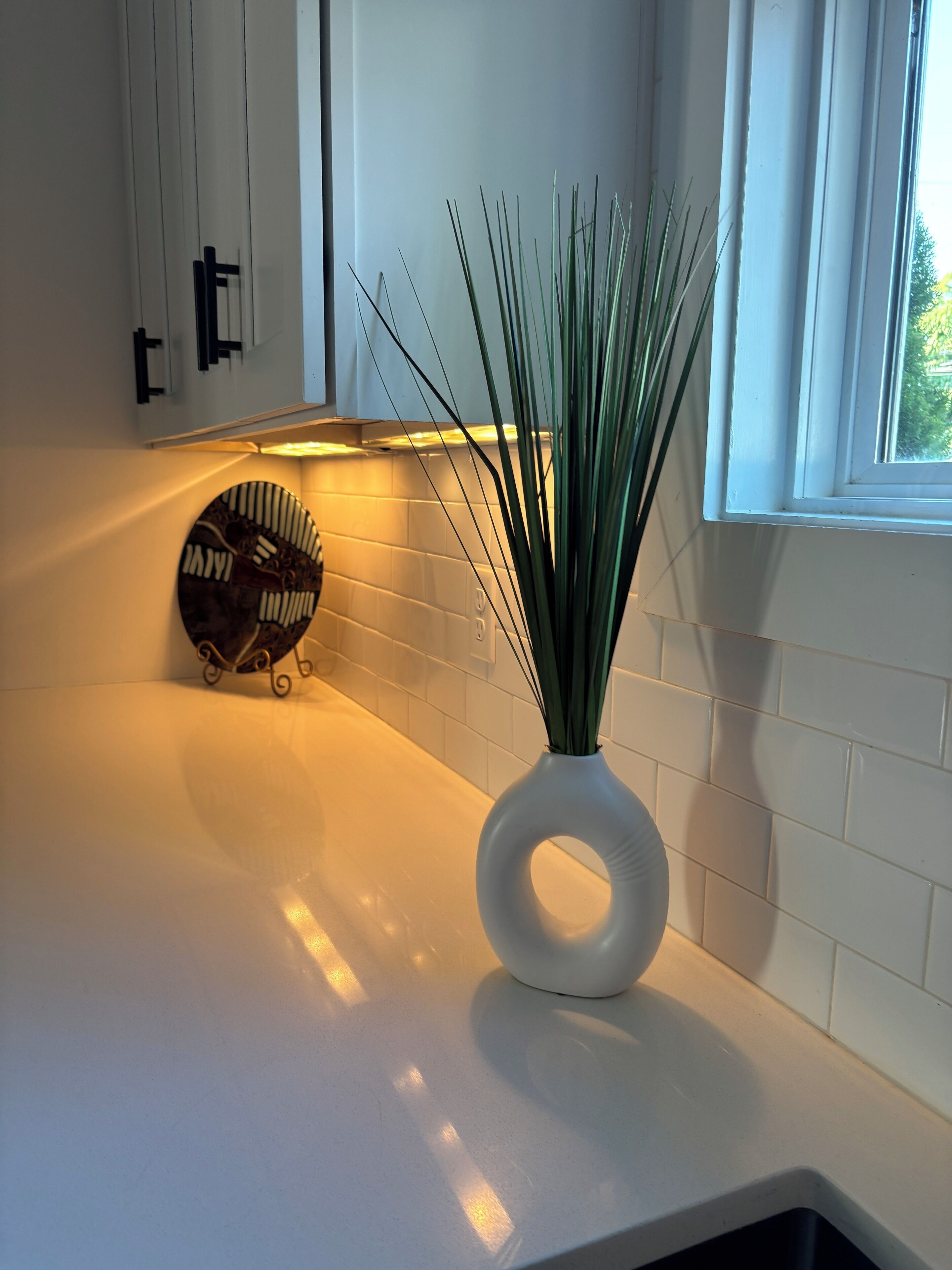Una cucina con mobili bianchi, un piano di lavoro bianco, un piatto decorativo e un vaso con piante verdi.
