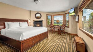 A hotel room with a large bed, a fireplace, a dining area with a table and chairs, a TV, and a window with a view. - Duck Inn Lodge (Whitefish)