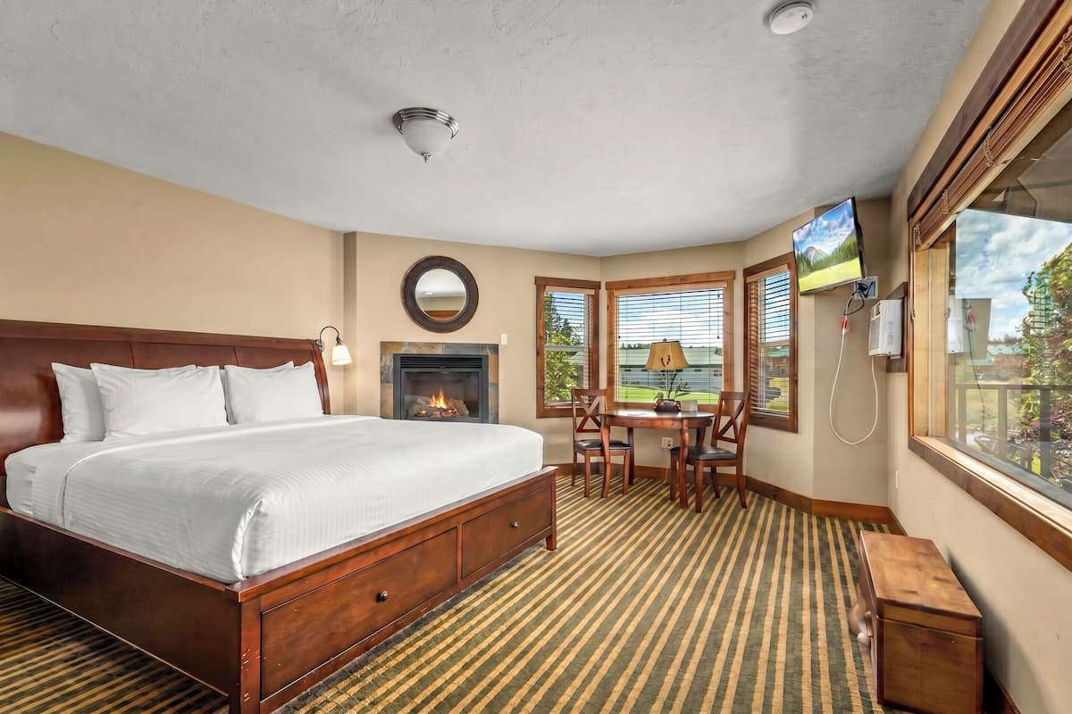 A hotel room with a large bed, a fireplace, a dining area with a table and chairs, a TV, and a window with a view.