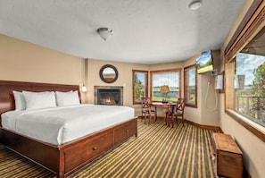 A hotel room with a large bed, a fireplace, a dining area with a table and chairs, a TV, and a window with a view. - Duck Inn Lodge (Whitefish)