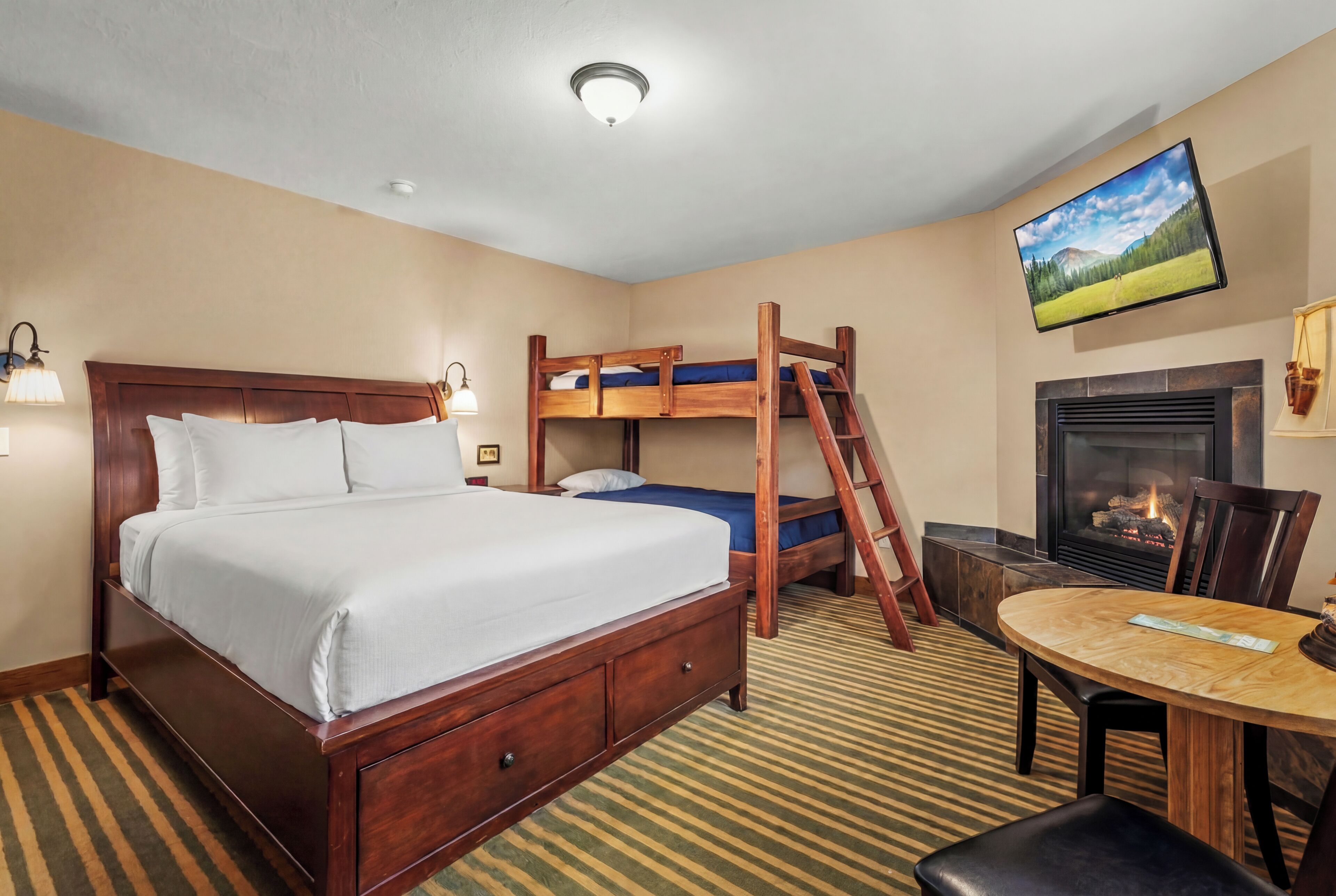 A hotel room with a large bed, a bunk bed, a fireplace, a TV, and a dining table.