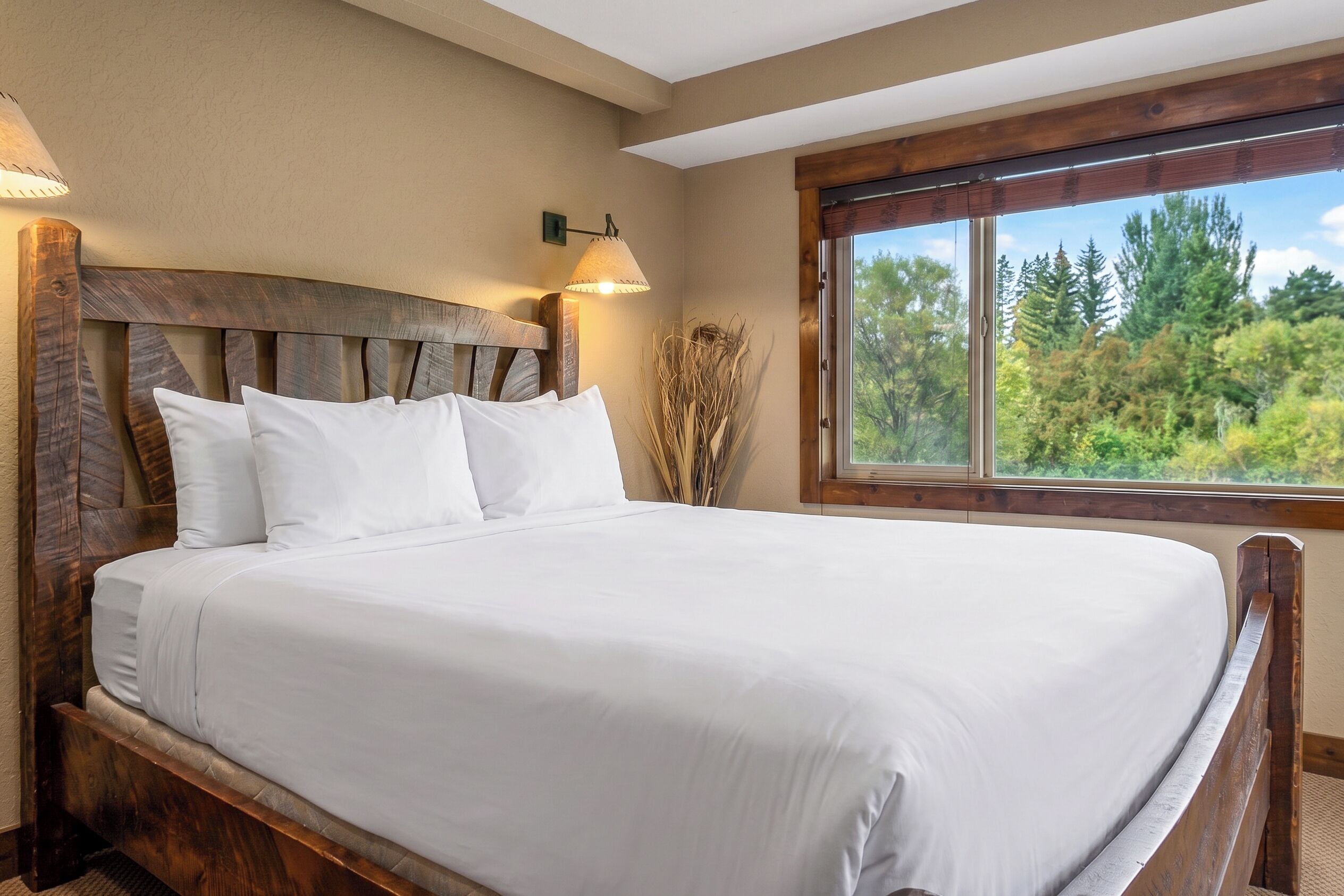 Una cama grande con sábanas blancas, cabecera de madera, lámpara fijada a la pared y ventana con vistas a árboles.