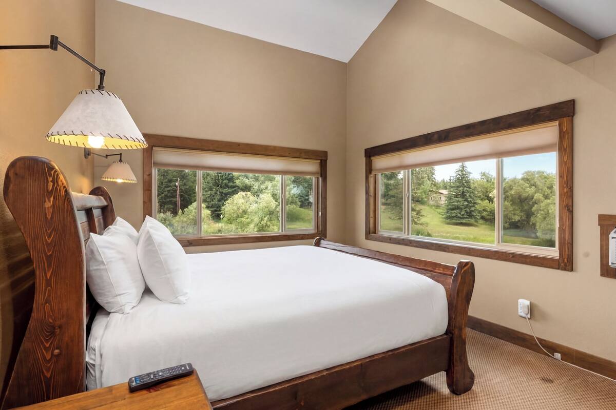 A bedroom with a large bed, two windows, a lamp, and a remote control on the nightstand.