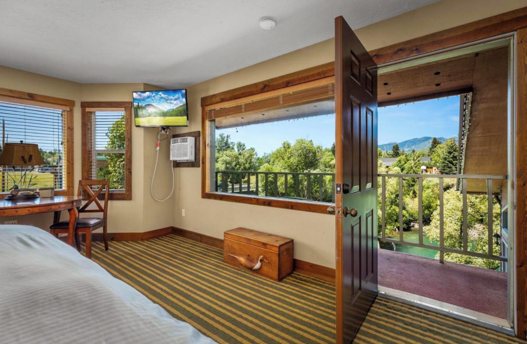 A room with a bed, a desk, a television, a window with a view of trees and mountains, and a door leading to an outdoor balcony.
