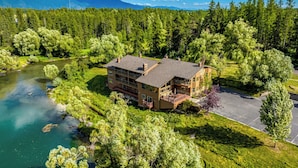 A multi-story lodge with a river view, surrounded by dense forest and mountains in the background. - Duck Inn Lodge (Whitefish)