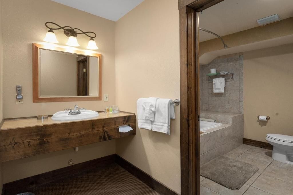A bathroom with a wooden vanity, a large mirror with lights, a bathtub, and a toilet.
