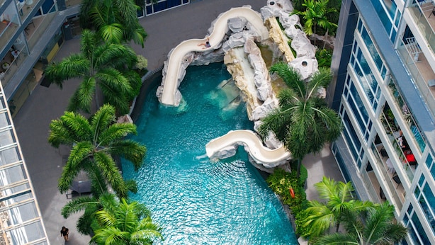 Une piscine intérieure avec un toboggan, entourée de palmiers et bordée d’une architecture moderne.