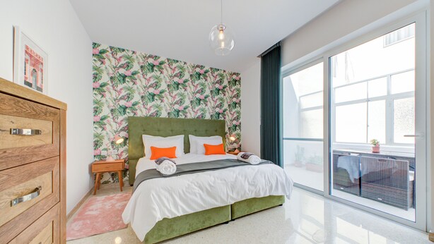 A modern bedroom with a green upholstered bed, white bedding, and orange pillows. There is a wooden dresser with drawers, a small bedside table with a lamp, and a large window with a view of the outdoors. - Colourful Spacious Maisonette with Yard (St. Paul's Bay)