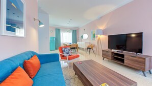 A modern living room with a blue sofa, orange cushions, a wooden coffee table, a TV, and a dining area with a table and chairs. - Colourful Spacious Maisonette with Yard (St. Paul's Bay)