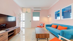 A modern living room with a flat-screen TV, wooden entertainment unit, blue sofa with orange cushions, and a coffee table. - Colourful Spacious Maisonette with Yard (St. Paul's Bay)